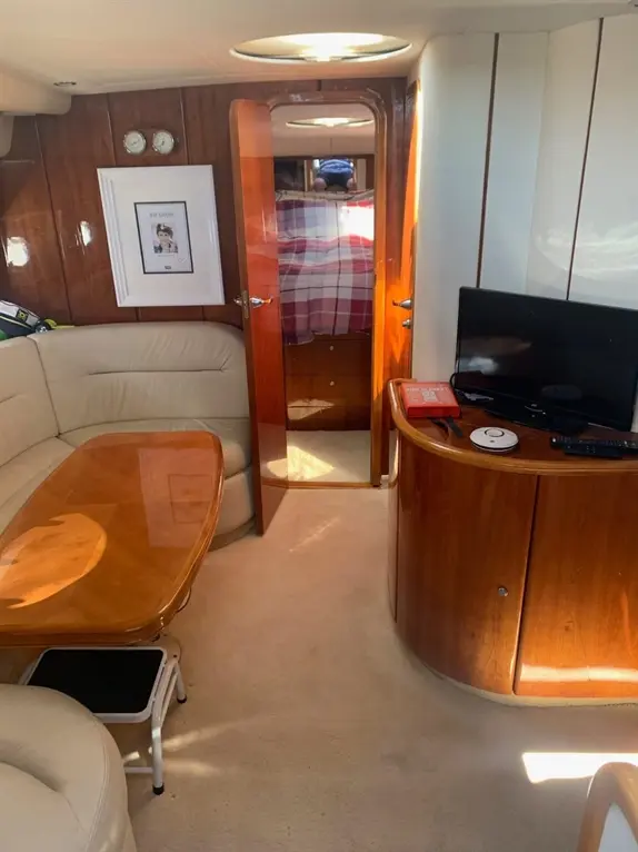 Interior of a yacht, featuring a leather sofa, wooden table, television, and an open door leading to a bedroom area.